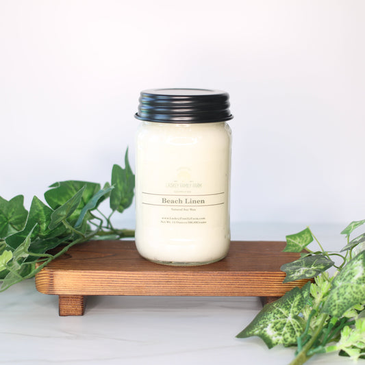 White candle with black lid on a wooden stand surrounded by green leaves against a white background
